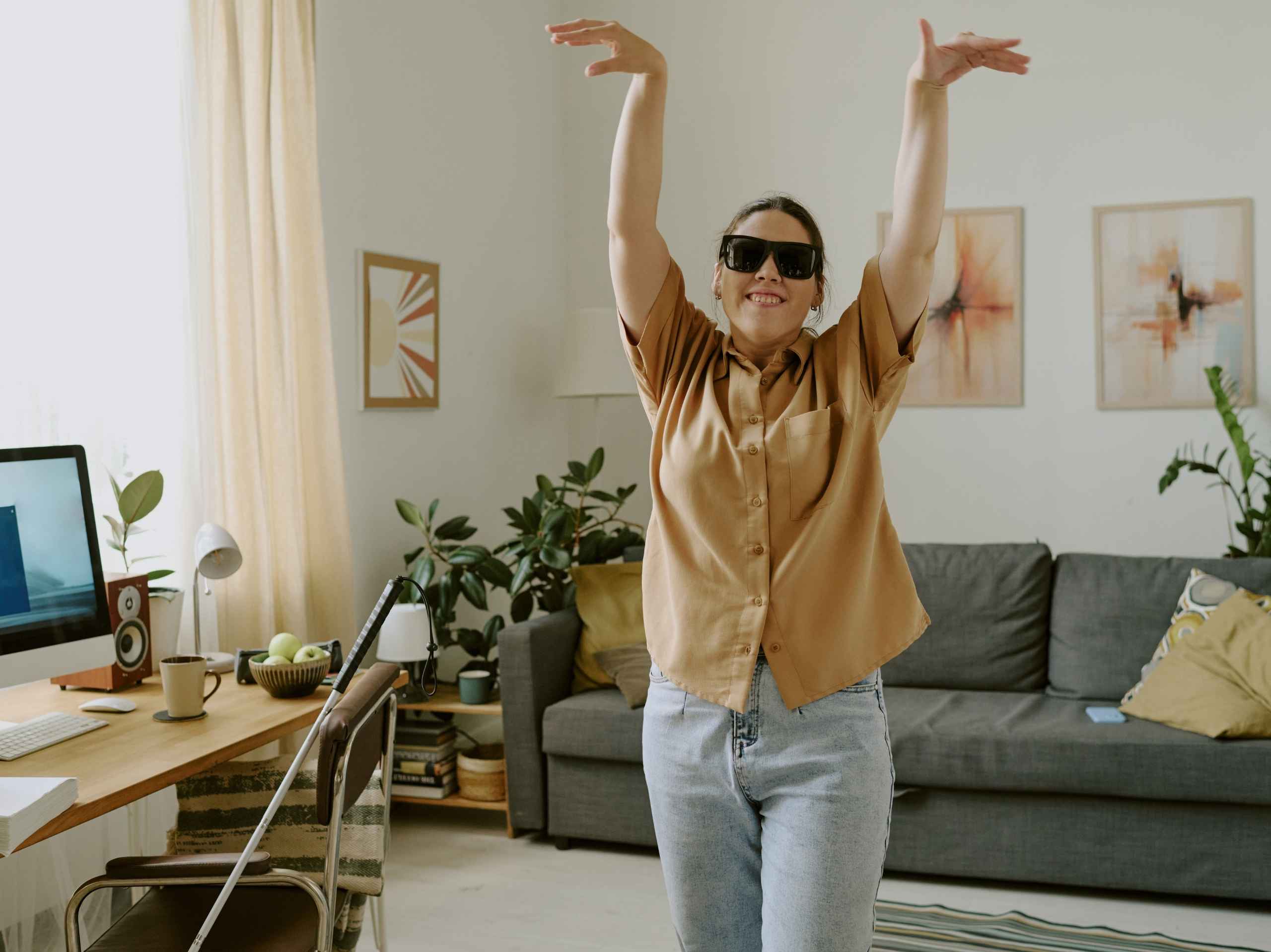 Smiling blind woman wearing sunglasses, raising her arms joyfully in a well-lit living room.