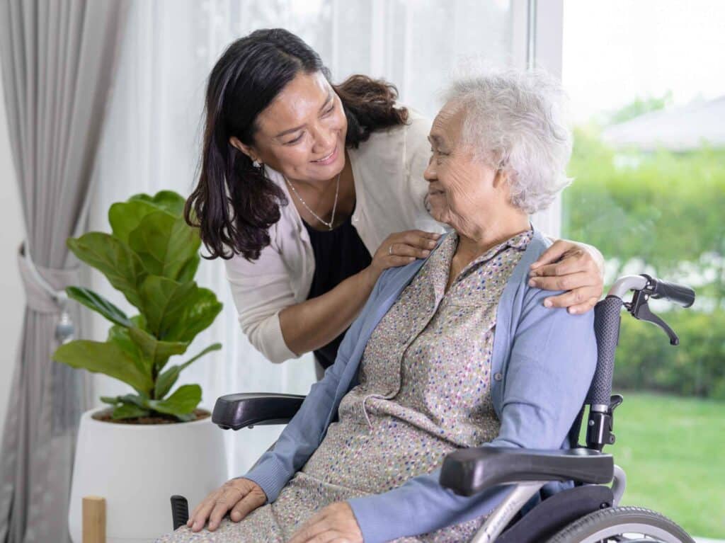 Female carer smiling and leaning in to support an elderly woman in a wheelchair indoors.