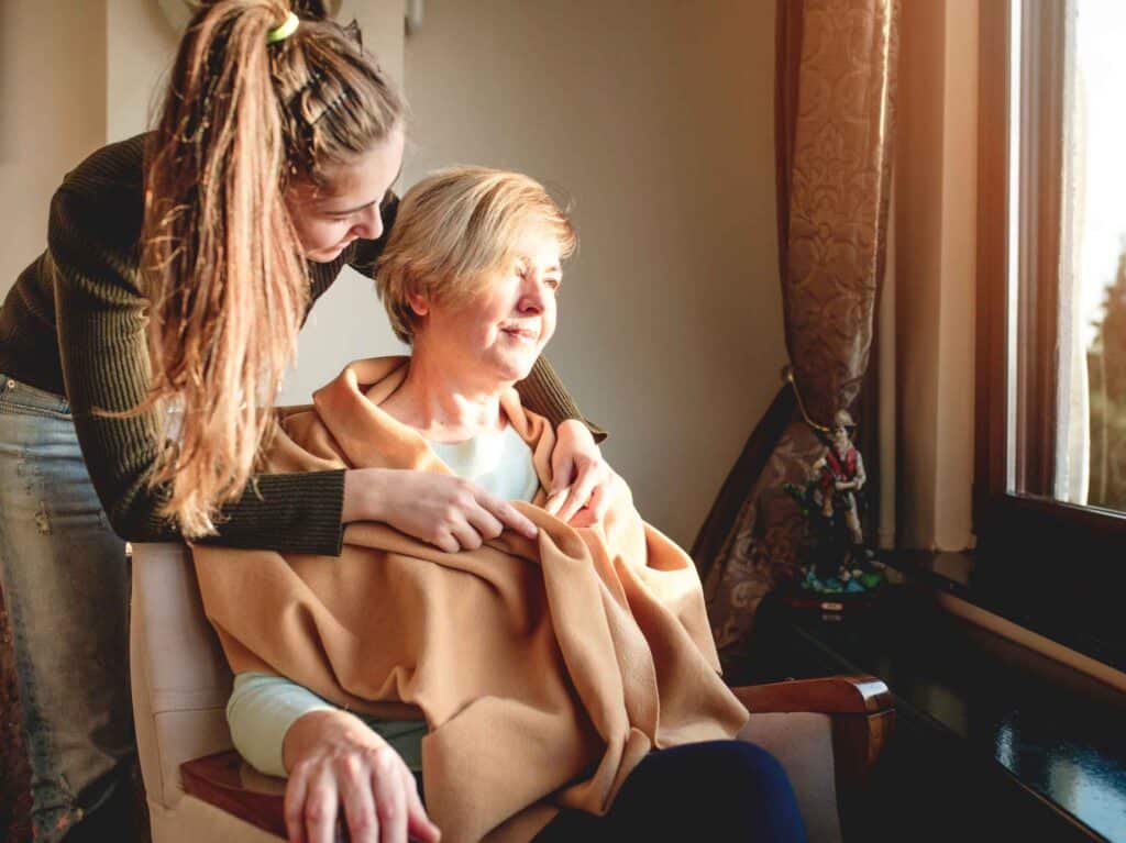 Young woman placing a warm blanket over an elderly lady seated by the window.