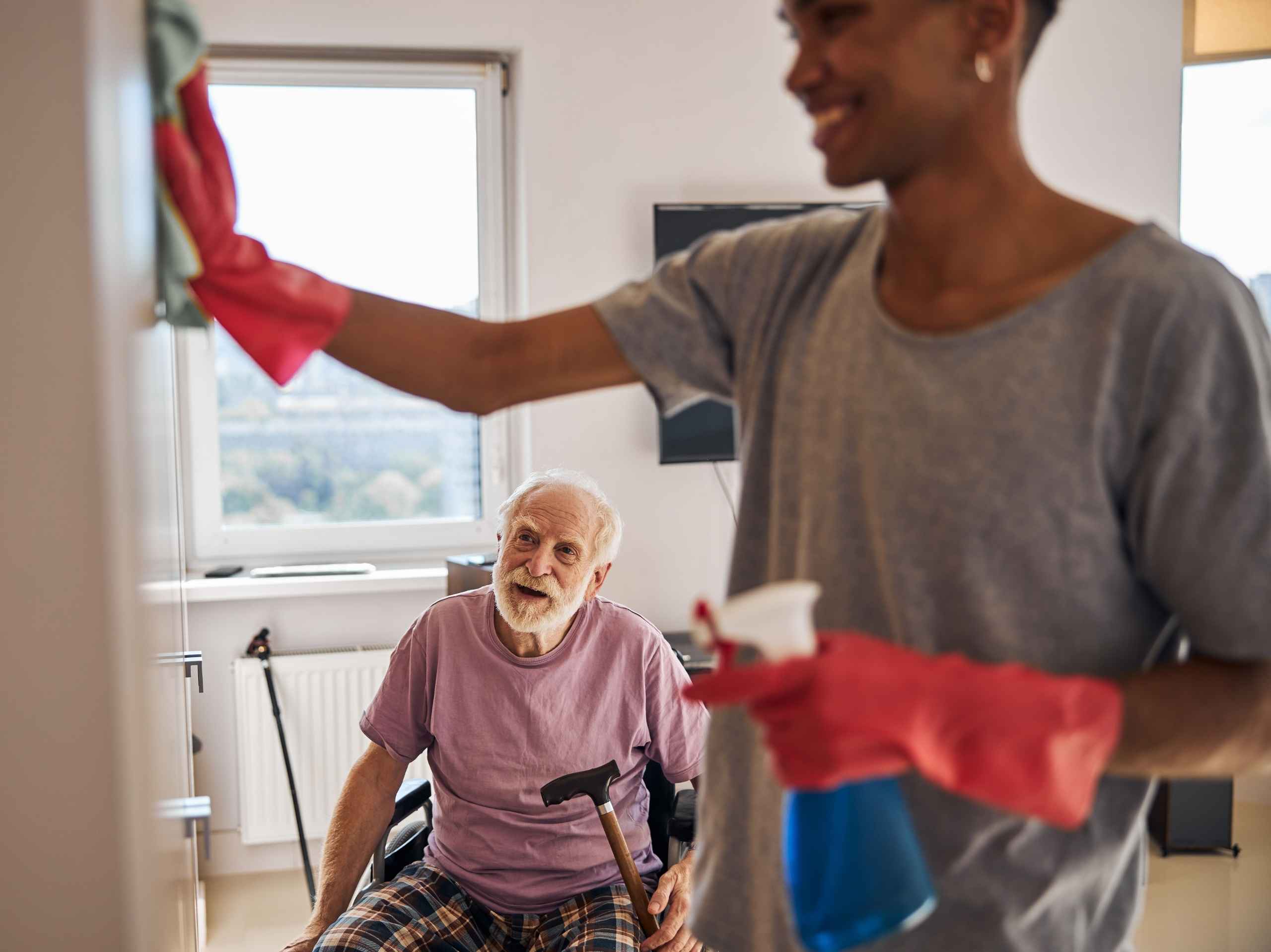 Elderly man smiling while a support worker cleans the room using gloves and a spray bottle.