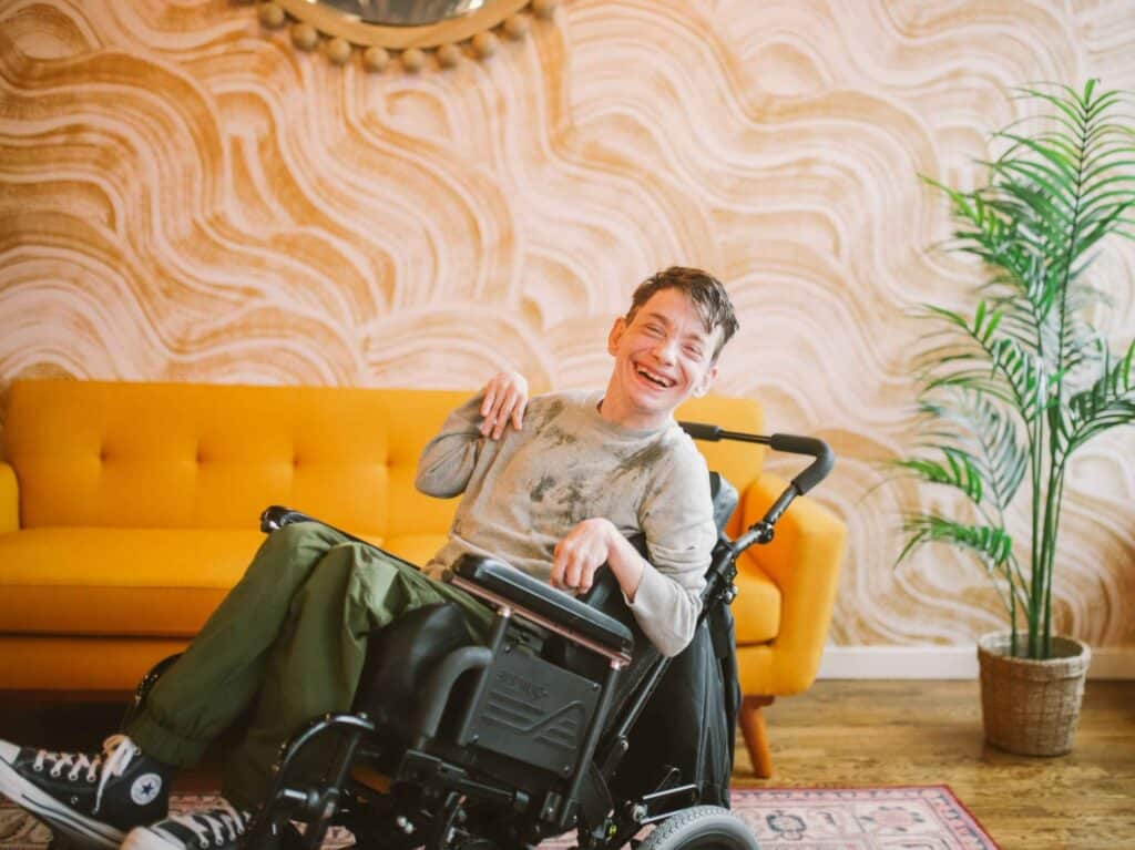 Young man in a wheelchair laughing joyfully in a colourful lounge room with a yellow couch.