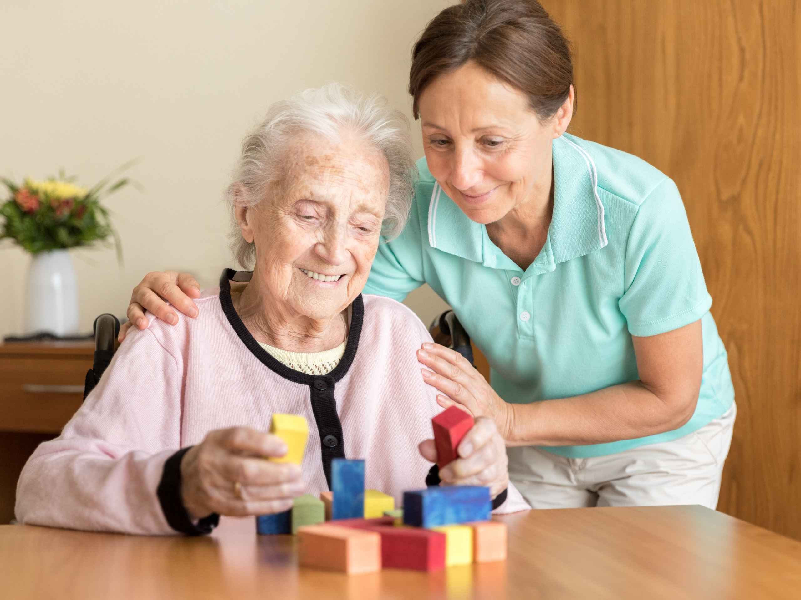 Senior woman smiling while playing with colourful blocks, assisted by a caring support worker.