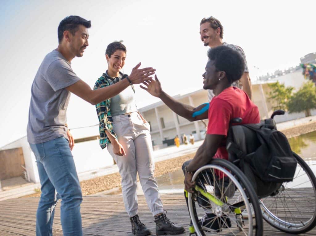 Man in wheelchair giving a high-five to a standing friend, surrounded by a happy group outdoors.