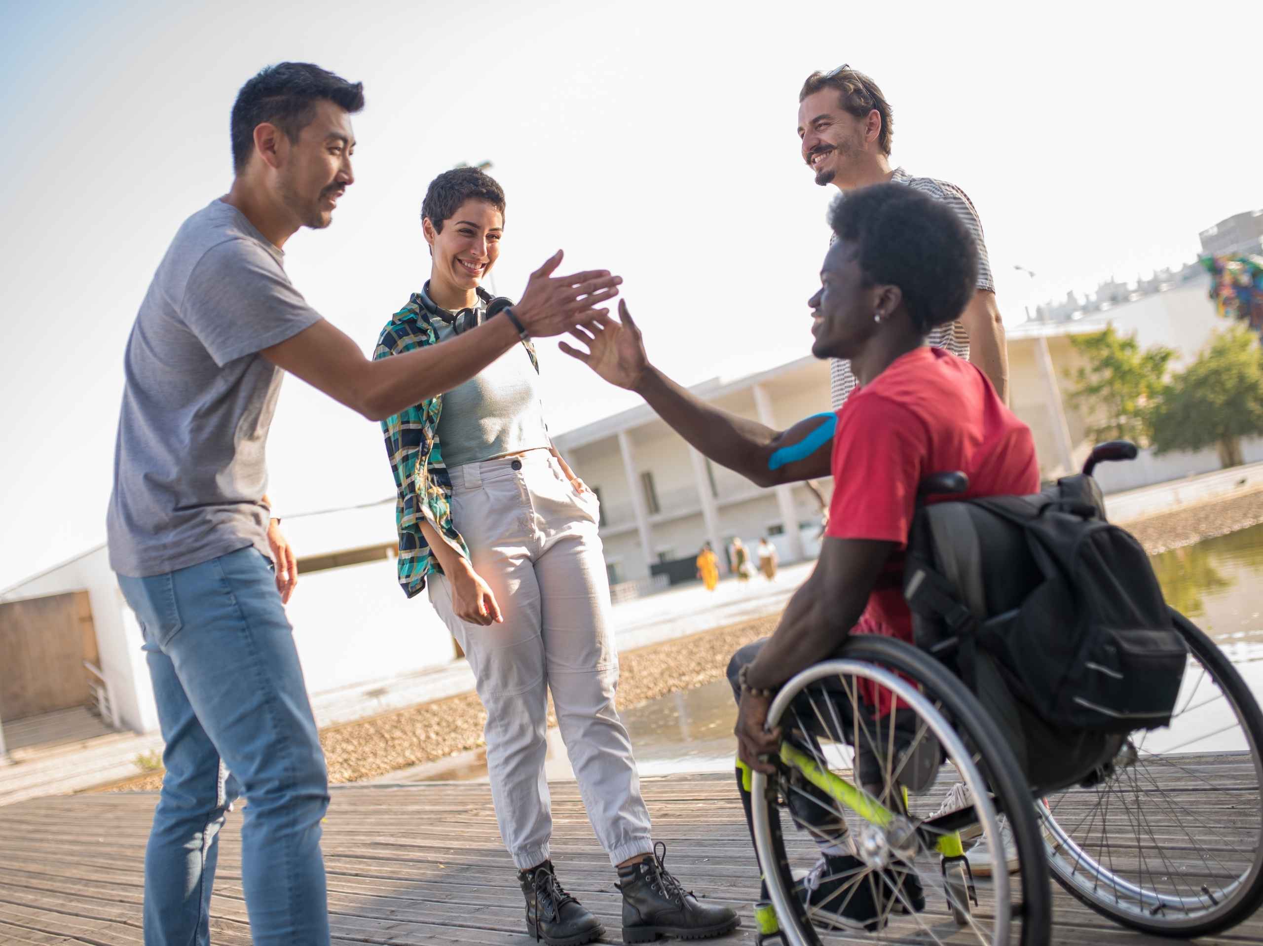 Man in wheelchair giving a high-five to a standing friend, surrounded by a happy group outdoors.
