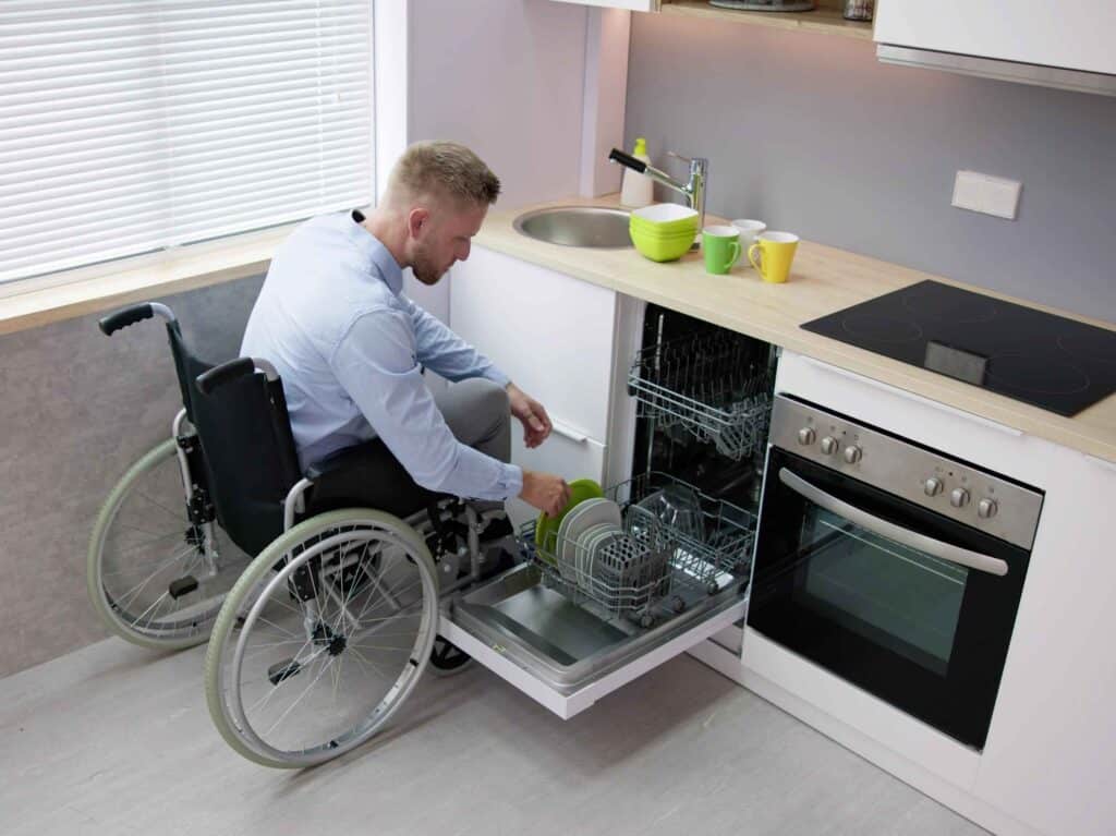 Man in a wheelchair loading plates into a dishwasher in a modern, accessible kitchen setup.