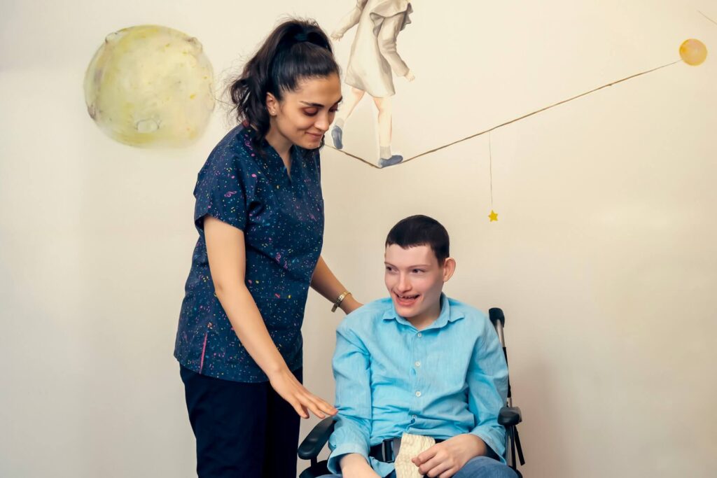 A caregiver smiling while assisting a child in a wheelchair.