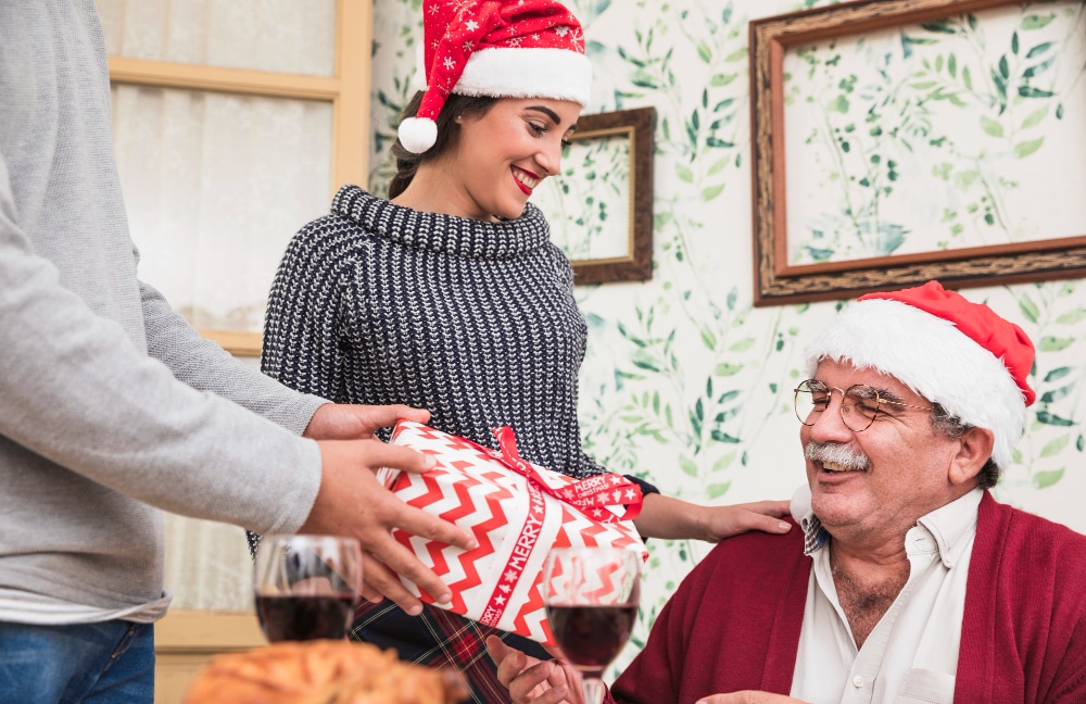 A joyful Christmas gift exchange between a young woman and an older man, both wearing Santa hats.
