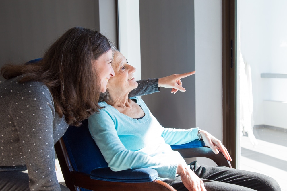 An elderly woman and her caregiver sharing a joyful moment while looking outside.